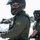 CBP Support to Super Bowl LIV Two U.S. Border Patrol agents are visible in an outdoor coastal or waterfront setting. The foreground agent is wearing a full-face motorcycle helmet, sunglasses, tactical vest, and green uniform with a clearly visible 'U.S. Border Patrol' shoulder patch. A second agent is visible in the background wearing similar gear including a helmet with goggles.