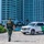 CBP Support to Super Bowl LIV A U.S. Border Patrol officer is walking on a sandy beach toward a white and green Border Patrol SUV (Chevrolet Tahoe). The scene takes place in front of a large modern high-rise building, suggesting a coastal urban environment. The officer is in full uniform with a tactical vest marked 'POLICE - U.S. Border Patrol'.