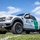 CBP Support to Super Bowl LIV A white U.S. Customs and Border Protection Ford F-150 Raptor truck is parked on a grass field under a partly cloudy blue sky. The vehicle is marked as a 'Mobile Command' unit with the DHS seal and green stripe characteristic of Border Patrol livery. The low-angle photograph emphasizes the truck's large off-road tires and imposing stance.