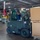 A male operator is driving an olive-green military-style Hyster 40 forklift inside a large industrial warehouse. The forklift is carrying two large cardboard boxes stacked on a wooden pallet. The warehouse environment is well-lit with overhead fluorescent lighting and features exposed ceiling infrastructure.