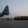 A U.S. Air Force C-130 Hercules transport aircraft (tail number 55828, assigned to AMC, nicknamed 'The Rock') is positioned on a flight line during hazy dawn or dusk conditions. Military personnel are gathered near the rear of the aircraft alongside a flatbed cargo loader vehicle stacked with palletized cargo. The scene depicts a cargo loading or unloading operation at a military airfield.