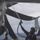 A military maintenance crew member is washing/hosing down the rotor blades of a helicopter on what appears to be a naval vessel's flight deck at sea. The person is using a pressurized hose to spray water upward onto the large dark rotor blades. The overcast sky and visible ocean horizon confirm an open-sea shipboard environment.
