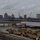 A military port terminal facility showing a large staging area with numerous tan/desert-colored military vehicles and equipment awaiting loading or offloading. Shipping containers are stacked near a terminal building with visible text 'SHIPPING TERMINAL'. A city skyline with cranes and a large gray military or cargo vessel is visible across the waterway in the background.