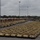 A large staging or marshaling yard contains hundreds of desert-tan painted military armored fighting vehicles, predominantly appearing to be M2/M3 Bradley Infantry Fighting Vehicles, arranged in dense rows. The vehicles are parked in an organized grid pattern at what appears to be a military port or rail facility, with freight rail cars visible in the background. The overcast sky and leafless trees suggest a late autumn or early spring setting in a temperate region.