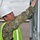 A U.S. Army soldier wearing OCP (Operational Camouflage Pattern) uniform is conducting an inspection at what appears to be a port or dock facility. The soldier is equipped with a white hard hat and high-visibility safety vest, suggesting a safety-compliant work environment. They are closely examining a metal structure, likely a shipping container or similar equipment.