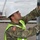 A U.S. Army soldier wearing OCP (Operational Camouflage Pattern) uniform with Airborne and 82nd Airborne Division patches is inspecting or working on a metal structure at what appears to be a military port or naval facility. The soldier is equipped with a white hard hat and high-visibility safety vest, indicating compliance with port safety regulations. A naval vessel and crane infrastructure are visible in the background.