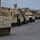A line of desert-tan painted U.S. Army armored tracked vehicles is staged in a large paved lot, likely a military staging or port facility. The vehicles appear to be Bradley Fighting Vehicles and M88 Recovery Vehicles, consistent with pre-deployment or equipment transfer operations. A single worker in a high-visibility vest and hard hat is visible near the vehicles in the background.