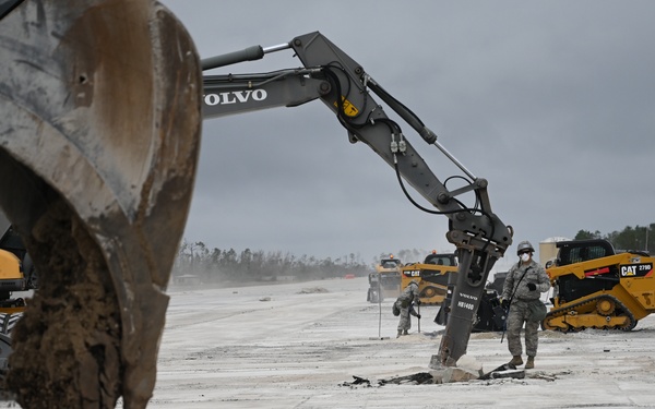 Military personnel in uniform are conducting airfield damage repair operations on what appears to be a concrete runway or taxiway. Multiple pieces of heavy construction equipment, including Volvo excavators and a Caterpillar tracked skid steer, are actively breaking up and removing damaged pavement sections. The overcast sky and stripped/damaged surface suggest post-disaster or training recovery operations.
