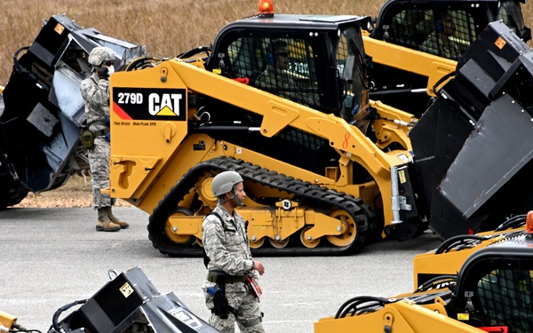 Military personnel in camouflage uniforms are seen among a fleet of yellow Caterpillar 279D compact track loaders equipped with rotary cutter/mulcher attachments. The machines are arranged in a staging area, likely on a military installation, with soldiers appearing to inspect or manage the equipment. This appears to be a military engineering or construction unit preparing for or conducting a field operation.