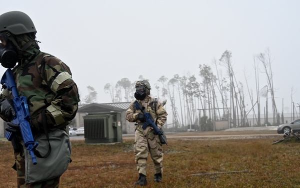 Two military personnel in full CBRN (Chemical, Biological, Radiological, Nuclear) protective gear, including gas masks and combat uniforms, are standing on a damaged military installation. Both carry blue-barreled training rifles, indicating this is likely a readiness exercise or drill. The surrounding environment shows significant storm damage, with downed trees, debris, and damaged structures visible in the foggy background.