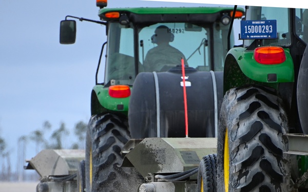A green John Deere tractor with the license plate '15D00293' marked 'ON BASE ONLY' is operating on what appears to be a military airfield or runway. The tractor is pulling a wide sweeper attachment with rotating brushes that are actively cleaning debris and dirt from the paved surface. Palm trees are visible in the background, suggesting a coastal or southern U.S. military installation.