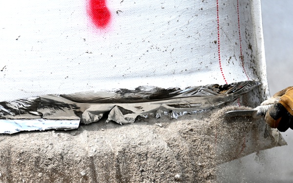 A close-up image showing a gloved hand using a tool to scrape or work a concrete or mortar surface. The surface appears wet or freshly applied, with material particles visibly dispersing in the air. A white fabric or tarp with red markings and metallic foil material is visible along the upper edge of the surface.