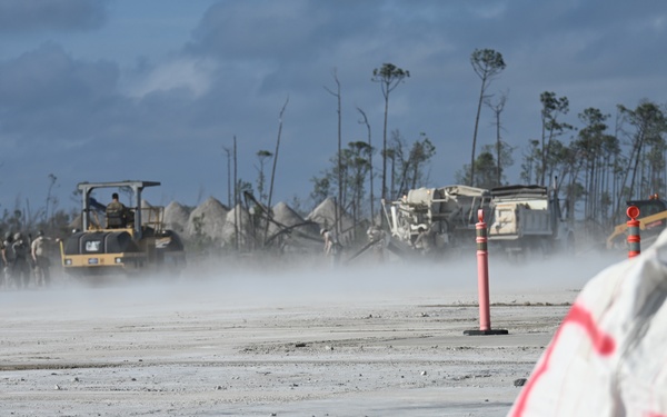 The image depicts an active construction or disaster recovery operation on a heavily damaged site, likely an airfield or road surface. Heavy equipment including a CAT roller/compactor is visible alongside personnel working in dusty conditions. Stripped, damaged trees in the background suggest prior severe storm or hurricane damage.