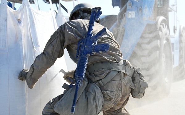 A soldier in full combat gear and camouflage uniform is crouching or taking cover behind large white bulk bags (FIBC/super sacks) in a dusty outdoor environment. The weapon carried appears to be painted blue, indicating this is likely a training exercise using simulated/inert weapons. Dust is being kicked up around the soldier, suggesting rapid movement near what appears to be a large vehicle or heavy equipment.