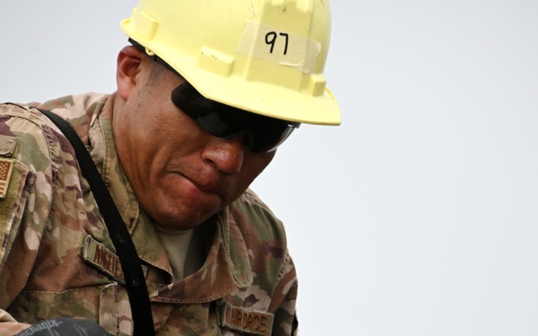 A U.S. Air Force service member wearing OCP (Operational Camouflage Pattern) uniform is engaged in construction or repair work, handling a piece of asphalt or concrete material. The individual is equipped with a yellow hard hat marked '97', tactical gloves, and sunglasses, indicating a field engineering or civil engineering task. The work appears to involve airfield or infrastructure repair operations.
