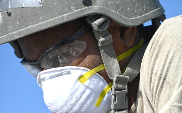 A worker is photographed in close-up wearing personal protective equipment including a hard hat, safety goggles, and an N95-style respirator mask. The individual appears to be engaged in dusty or hazardous work, as evidenced by white dust/powder coating their clothing, goggles, and skin. The image is taken against a clear blue sky background.