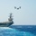 A large naval aircraft carrier is underway at sea under clear blue skies, with a tiltrotor aircraft flying overhead. The carrier's flight deck is visible with what appear to be parked aircraft, and the ship is creating a white wake as it moves through deep blue ocean water. The scene depicts active naval operations in open ocean conditions.