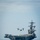 A U.S. Navy aircraft carrier, visibly identified as the USS Theodore Roosevelt (CVN-71), is underway at sea in open blue ocean waters. An MV-22 Osprey tiltrotor aircraft is approaching or departing the flight deck. The American flag is flying from the island superstructure, and multiple aircraft are visible on the flight deck.