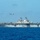 A large US Navy amphibious assault ship is underway on open blue ocean under clear skies. The flight deck is populated with multiple aircraft, and at least one helicopter is airborne above the vessel. The ship appears to be conducting flight operations or transit operations.