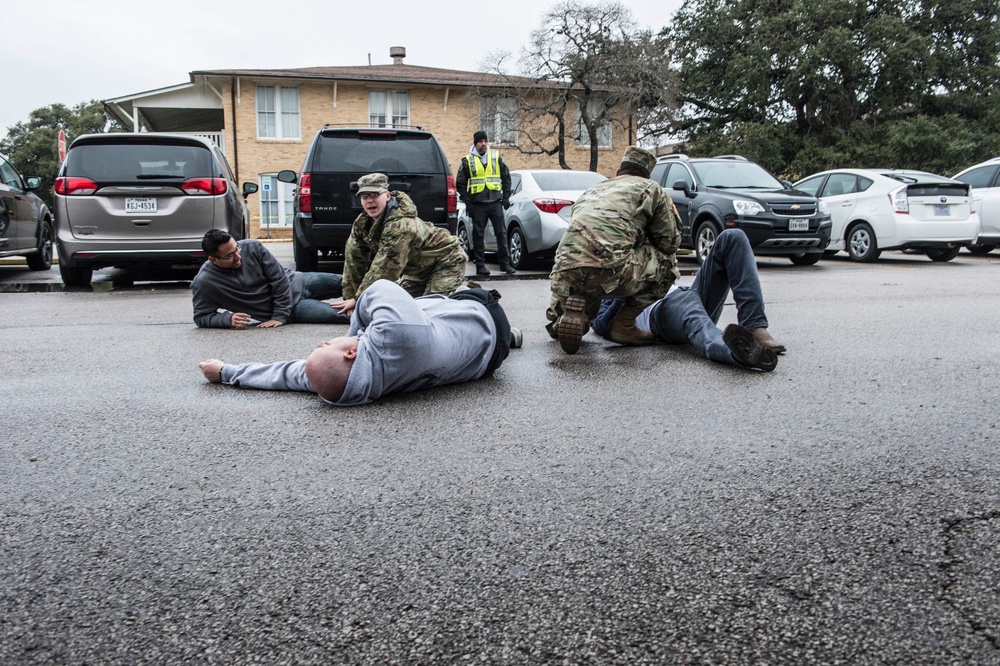 The image depicts what appears to be a military or law enforcement training exercise in a parking lot. Personnel in camouflage uniforms are detaining or assisting civilians on the ground, while a safety observer in a high-visibility vest watches from the background. The setting includes a brick building, multiple parked vehicles, and bare winter trees.