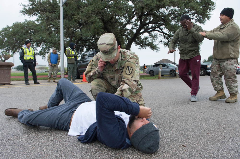 The image depicts what appears to be a military training exercise or drill taking place in a parking lot. A soldier in OCP (Operational Camouflage Pattern) uniform is kneeling over a person lying on the ground, while two other individuals in civilian and military attire appear to be engaged in a simulated confrontation nearby. Several observers in high-visibility vests and civilian clothing watch from the background.