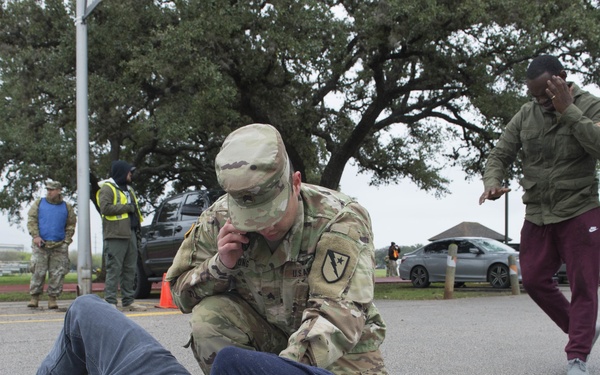 The image depicts what appears to be a military training exercise or drill taking place in a parking lot. A soldier in OCP (Operational Camouflage Pattern) uniform is kneeling over a person lying on the ground, while two other individuals in civilian and military attire appear to be engaged in a simulated confrontation nearby. Several observers in high-visibility vests and civilian clothing watch from the background.