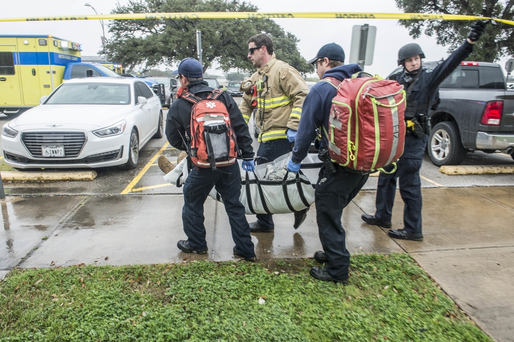 Emergency responders are carrying a patient on a stretcher or litter across a wet parking lot during what appears to be a mass casualty or active incident response. Personnel include EMS workers with medical backpacks, a firefighter in turnout gear, and a law enforcement officer in tactical gear lifting yellow crime scene tape. An ambulance and multiple vehicles are visible in the background.