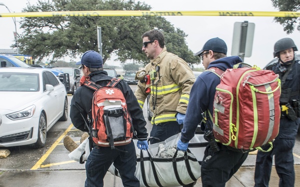 Emergency responders are carrying a patient on a stretcher or litter across a wet parking lot during what appears to be a mass casualty or active incident response. Personnel include EMS workers with medical backpacks, a firefighter in turnout gear, and a law enforcement officer in tactical gear lifting yellow crime scene tape. An ambulance and multiple vehicles are visible in the background.