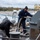 USNS Benavidez Arrives in Beaumont Two workers are handling mooring lines on the deck of a naval or commercial vessel docked at a port facility. One worker wearing a white hard hat is managing lines near a bollard, while the other in navy blue coveralls and a black beanie is feeding a thick rope into a mooring winch. The equipment and markings suggest this is a U.S. Navy or government vessel undergoing line-handling operations.