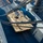 AGILE DEFENDER 2020 An aerial/overhead view shows a desert-tan M1 Abrams main battle tank being loaded or unloaded via a ship's ramp or gangway, with heavy steel cables visible in the foreground. The operation appears to be a military vehicle embarkation or debarkation at a port facility. At least one safety-vested worker is visible overseeing the operation from the upper deck area.