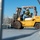 AGILE DEFENDER 2020 A worker in a yellow safety vest and white hard hat operates a yellow Caterpillar forklift outdoors under a clear blue sky. The forklift is carrying a wire mesh cage/container filled with materials, viewed through a metal grating in the foreground. The setting appears to be an industrial or construction yard with a concrete surface.