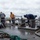 USNS Benavidez Arrives in Beaumont Several workers are on the wet deck of a vessel, handling mooring lines and ropes in overcast, damp conditions. The crew appears to be engaged in docking or undocking operations, managing heavy lines around deck cleats and bollards. The scene suggests a working waterfront environment with industrial maritime equipment visible.