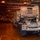 AGILE DEFENDER 2020 The image shows the interior vehicle deck of a military transport or amphibious ship, illuminated by warm overhead lighting. Multiple large military trucks and heavy equipment transporters are parked in rows within the enclosed cargo hold. The vehicles appear to be US Army equipment secured with cargo nets and tie-down chains.