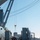 AGILE DEFENDER 2020 A port/dock loading operation is underway alongside a large gray vessel. A ship's crane is actively lifting a cargo container or unit load using a spreader bar and rigging cables. Multiple pieces of ground support equipment are visible on the concrete pier.