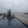 USNS Benavidez Arrives in Beaumont An aerial view from the elevated position of a large military or government vessel navigating a wide, muddy waterway under overcast, rainy conditions. The ship's stern deck is prominently visible, featuring a clearly marked helicopter landing pad (helipad) with an 'H' symbol. A cable-stayed or arch bridge is visible in the misty background, suggesting the vessel is transiting through an industrial port or river channel.