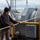 USNS Benavidez Arrives in Beaumont A male worker is handling mooring lines on the deck of a large vessel at a port or industrial waterfront facility. He is wearing cold-weather work gear and gloves while managing thick braided ropes connected to large gray winch drums. The background reveals an industrial port environment with cranes, buildings, and overcast skies.