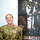 A U.S. Navy service member named Seale, wearing Navy Working Uniform (NWU) digital camouflage, stands smiling in front of a framed vintage military recruitment poster. The poster visible in the background reads 'OVER THERE' and depicts a WWI-era sailor in white uniform, consistent with classic American wartime propaganda artwork. The setting appears to be an indoor military office or administrative space.