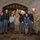 A group of approximately nine individuals pose for a formal group photograph inside what appears to be a military academy or institutional building with Gothic stone architecture. The group includes cadets in gray dress uniforms, active duty military personnel in camouflage (OCP/MultiCam) uniforms, and one civilian woman in casual attire. The setting and uniforms suggest this is an official or ceremonial occasion at a military institution.