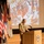 A military officer in multicam/camouflage uniform stands at a wooden podium delivering a presentation in what appears to be an auditorium or lecture hall. Behind him, a projected slide displays a historical battle painting with partially visible caption text reading 'EXP... CHINE, 186...' suggesting a topic related to historical military campaigns. Several military flags, including the US flag and a UK Union Jack, are displayed on the left side of the stage.