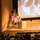 A military officer in uniform is delivering a presentation at a podium on an auditorium stage, with a large projected screen behind him showing two civilians seated together. The venue appears to be a formal military or government auditorium with multiple flags displayed on stage, including the US flag and what appears to be a UK flag. A large audience of what appear to be military personnel and civilians is seated in the auditorium.