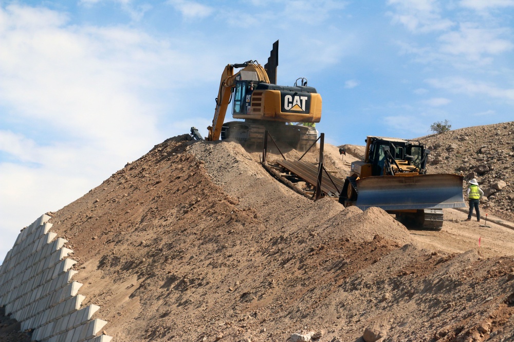Workers use a track hoe to pull a sled with barrier panels up a hill