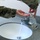 Two individuals wearing military camouflage uniforms are at a bathroom sink, with one person dispensing liquid soap or hand sanitizer from a pump bottle into the other person's outstretched hand. The water faucet is running, indicating an active handwashing procedure. The setting appears to be a military facility bathroom with a granite-style countertop.