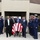 A military funeral procession is taking place outside what appears to be a funeral home or chapel. A flag-draped casket is being wheeled out by civilian pallbearers through a corridor of uniformed U.S. Coast Guard personnel rendering hand salutes. The scene depicts a formal military honors ceremony for a deceased service member.