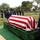 A military funeral ceremony is taking place at an outdoor cemetery, with uniformed personnel standing at attention around a flag-draped casket. The American flag is draped over the casket in the traditional manner, and a service member appears to be reading from a document, likely delivering remarks or a eulogy. The ceremony involves a large formation of military personnel in dress uniforms consistent with the United States Coast Guard.