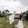 A military funeral ceremony is taking place at an outdoor cemetery. A flag-draped casket is positioned near a green canopy tent, surrounded by uniformed personnel saluting and civilian mourners. A formation of military personnel in blue uniforms stands at attention on the left side of the scene.