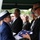 A military funeral ceremony is taking place outdoors, likely at a cemetery. A uniformed service member wearing white gloves is presenting a folded American flag to a civilian, while several mourners dressed in dark formal attire look on solemnly. The scene depicts the traditional flag presentation ritual conducted at military burial services.