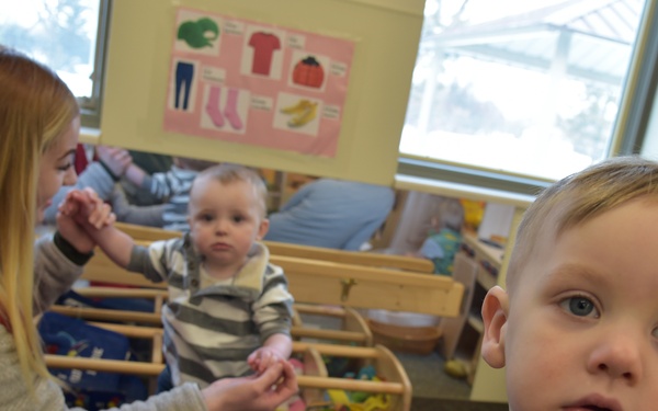 A daycare or early childhood classroom setting with young toddlers and a female caregiver. In the foreground, a toddler wearing a blue 'The Amazing Spider-Man' t-shirt looks toward the camera. In the background, a woman with long blonde hair kneels on the floor attending to another toddler in a striped shirt.