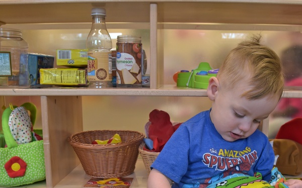 A toddler sits on a carpeted floor in front of a low wooden shelving unit, playing with a soft cloth book featuring a crocodile/alligator design. The child is wearing a blue 'The Amazing Spider-Man' t-shirt, gray sweatpants, and black Batman sneakers. The setting appears to be a daycare or preschool classroom environment.