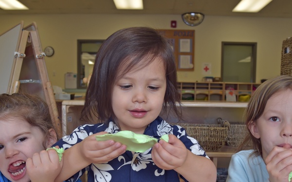 Three young children are seated at a table in what appears to be a preschool or daycare classroom, playing with green play dough. Each child is holding and manipulating pieces of the green modeling clay, with various expressions of engagement and playfulness. The classroom setting includes shelving units, cubbies, an easel, and other typical early childhood education furnishings in the background.