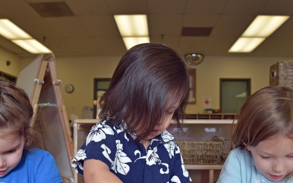 Three young children are seated at a table in what appears to be a preschool or daycare classroom, engaged in a play-dough activity. Each child is manipulating green play-dough using their hands and tools. The setting is a well-lit indoor classroom with shelving, an easel, and educational materials visible in the background.